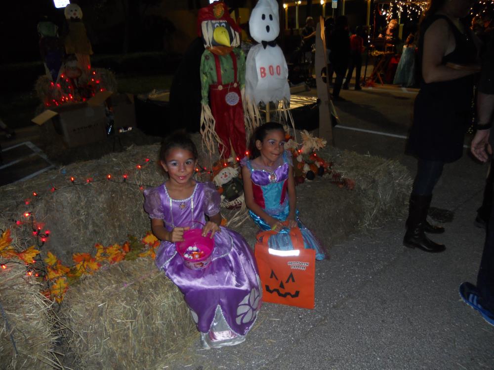 Girl in purple princess costume holding pink candy bucket sits on hay bales next with girl in blue princess outfit holding orange jack-o-lantern candy bag